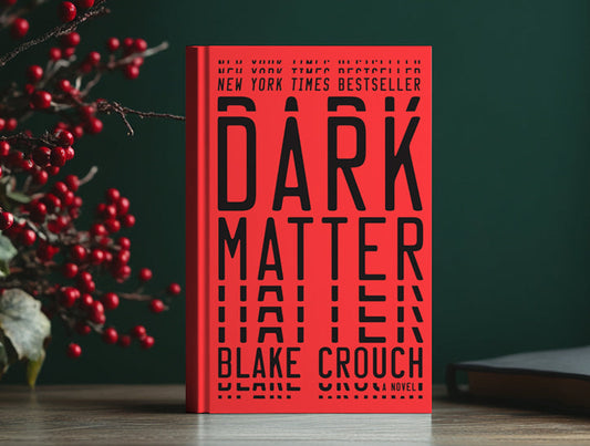 A hardcover copy of Dark Matter by Blake Crouch. The book is placed on a wooden surface with festive red berries and green foliage on the side, set against a dark green background.