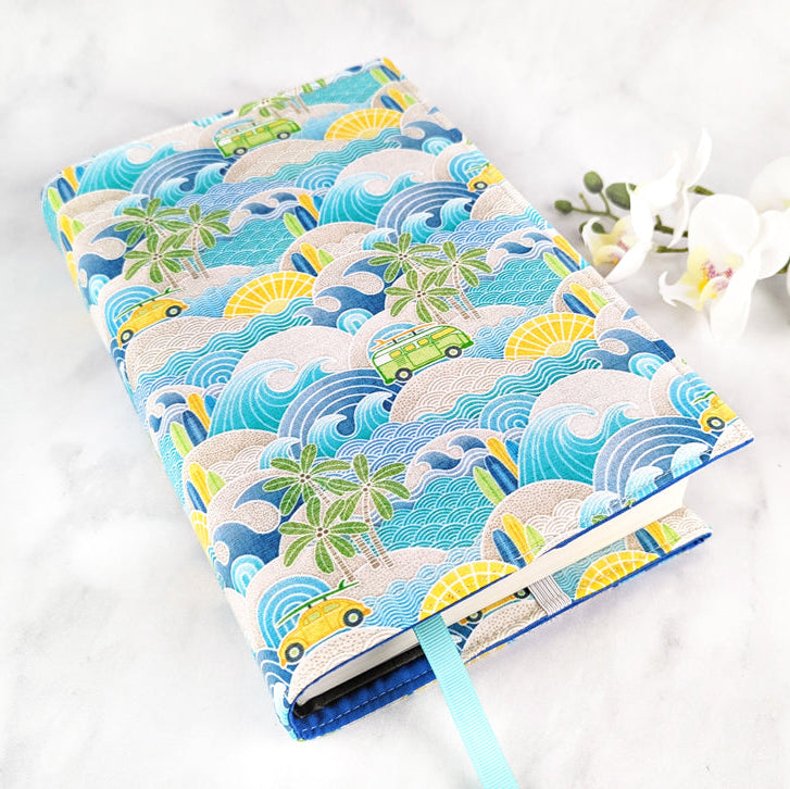 Fabric book cover with a vibrant beach scene featuring waves, palm trees, surfboards, yellow and green vans, and setting suns; styled with a blue ribbon bookmark and laid on a marble background with white orchids.
