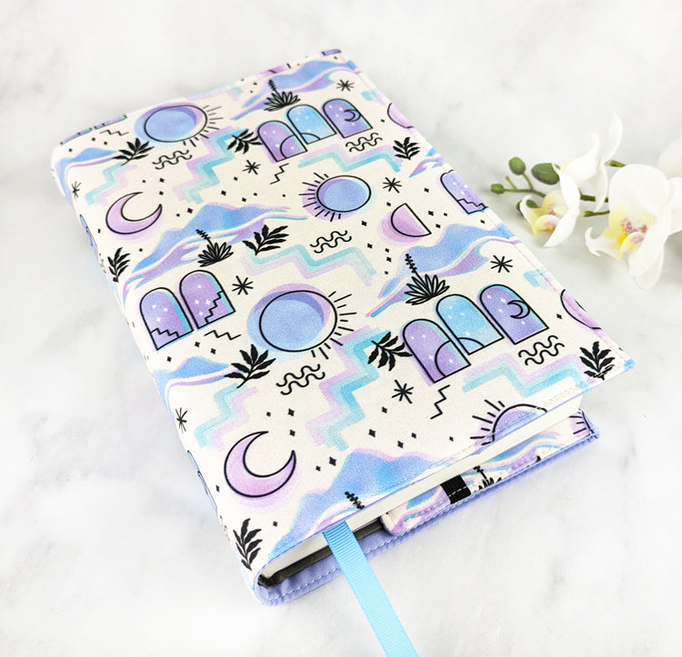 Fabric book cover with a pastel purple and blue celestial design featuring moons, suns, arches, and abstract shapes, fitted on a book with a light blue ribbon bookmark, displayed beside white flowers.