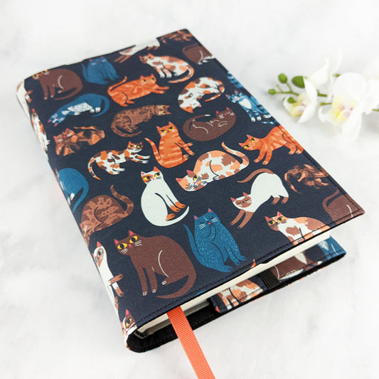 Fabric book cover featuring a playful pattern of illustrated cats in various colors and poses on a dark background, accented with an orange ribbon bookmark.