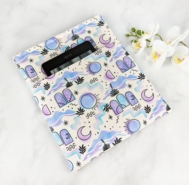 Fabric book sleeve with a pastel celestial pattern featuring moons, suns, stars, and abstract shapes, displayed with white flowers on a white surface.