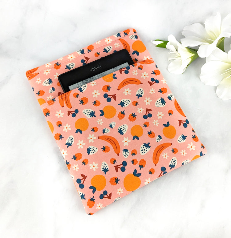 medium padded book sleeve with pocket. fabric showcases oranges, bananas, strawberries, blueberries and flowers on a light pink background.