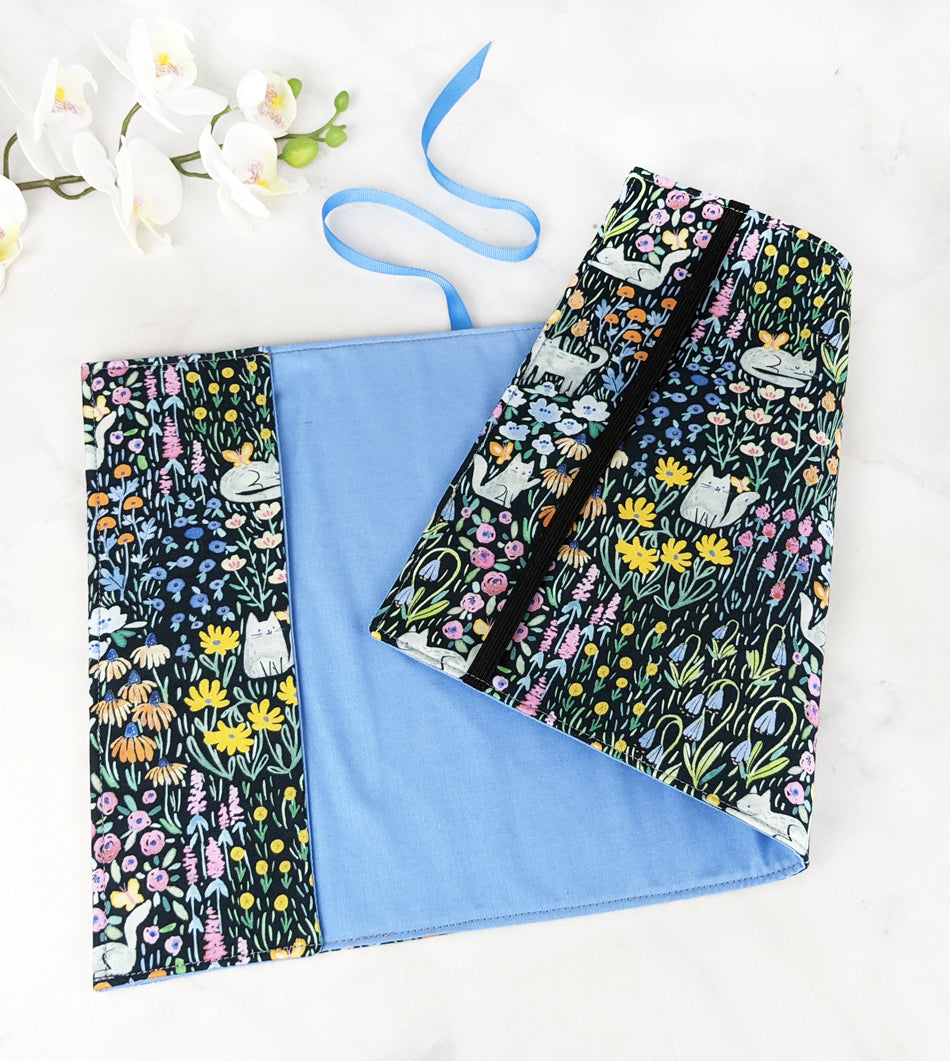 Fabric book cover featuring a whimsical print of white cats lounging and playing among colorful wildflowers on a black background, finished with a blue ribbon bookmark.
