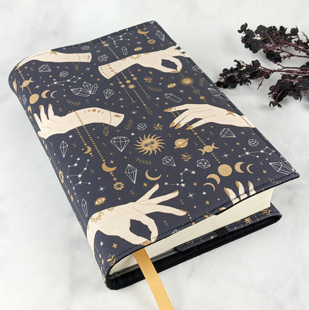 Fabric book cover with a celestial design featuring cream-colored hands, golden suns, moons, stars, constellations, and crystals on a dark navy background.