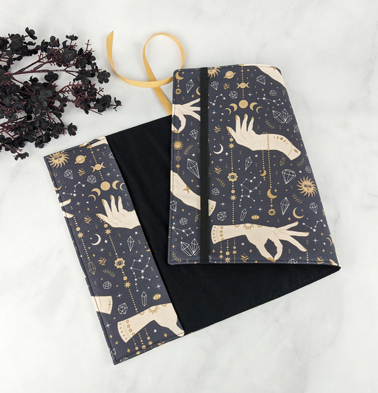 Fabric book cover laid open to show both exterior and interior. The outer fabric features a celestial design with mystical hands, moons, stars, and crystals in gold and cream on navy blue. Inside is solid black with a matching gold ribbon bookmark.