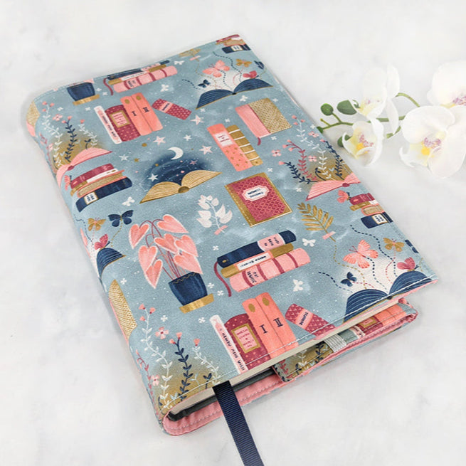 Fabric book cover with a dreamy book-themed design featuring illustrated books, open pages, potted plants, and moons in soft shades of pink, navy, and gold on a dusty blue background, finished with a dark navy ribbon bookmark.