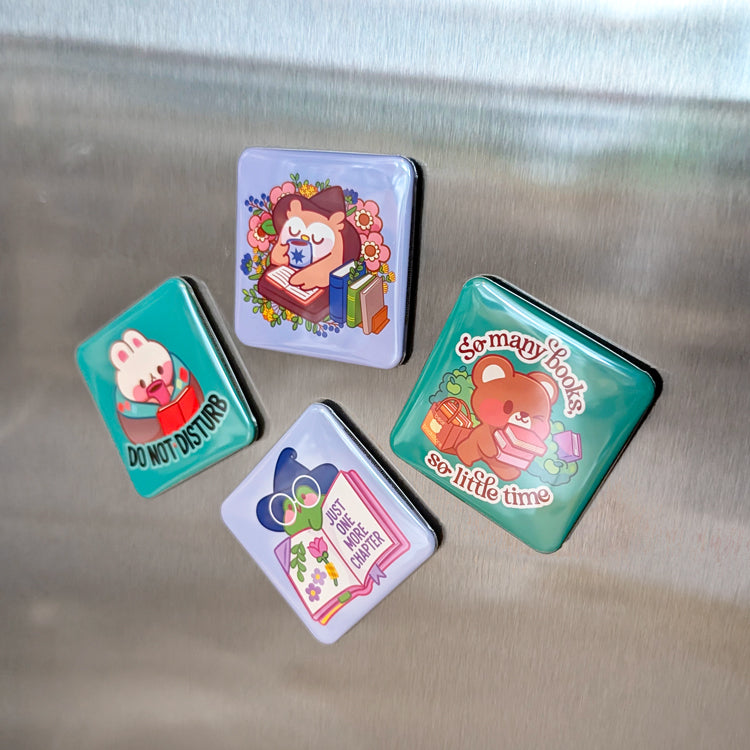 Four colorful square magnets with cute animal designs on a metallic surface. The magnets feature: a bunny reading with "DO NOT DISTURB," an owl reading a book surrounded by flowers, a bear carrying books with "So many books, so little time," and a frog in a witch hat with "JUST ONE MORE CHAPTER."