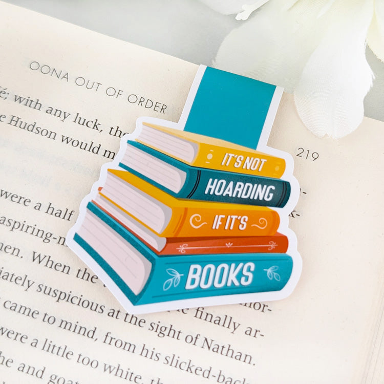 Magnetic bookmark featuring a colorful stack of books with the text 'It's not hoarding if it's books,' clipped to an open book.
