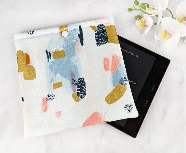 padded kindle oasis sleeve with snap. fabric showcases paint brush strokes in shades of navy blue, gold, pink and gray on a white background.