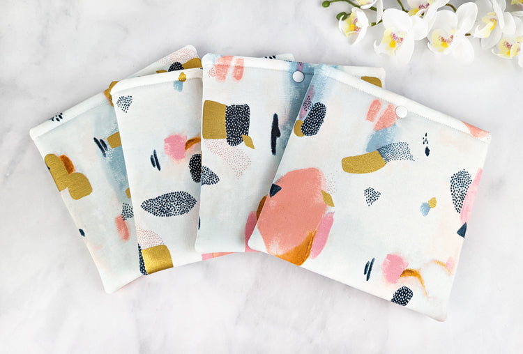 four padded kindle oasis sleeves with snap. fabric showcases paint brush strokes in shades of navy blue, gold, pink and gray on a white background.