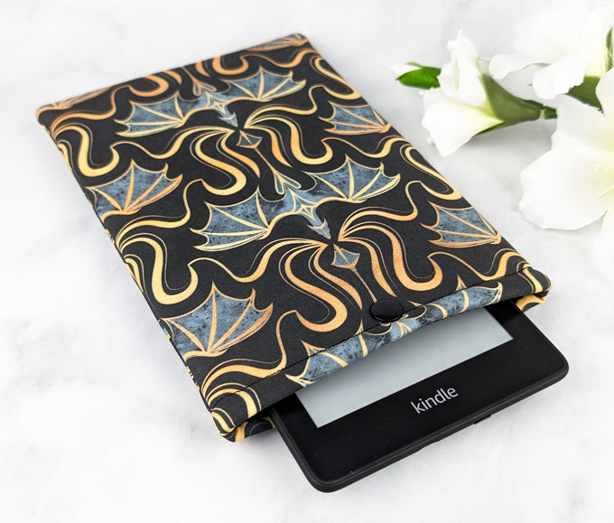 Padded Kindle Paperwhite sleeve with a bold pattern of gold and gray dragons on a black background, displayed with white flowers on a white surface.