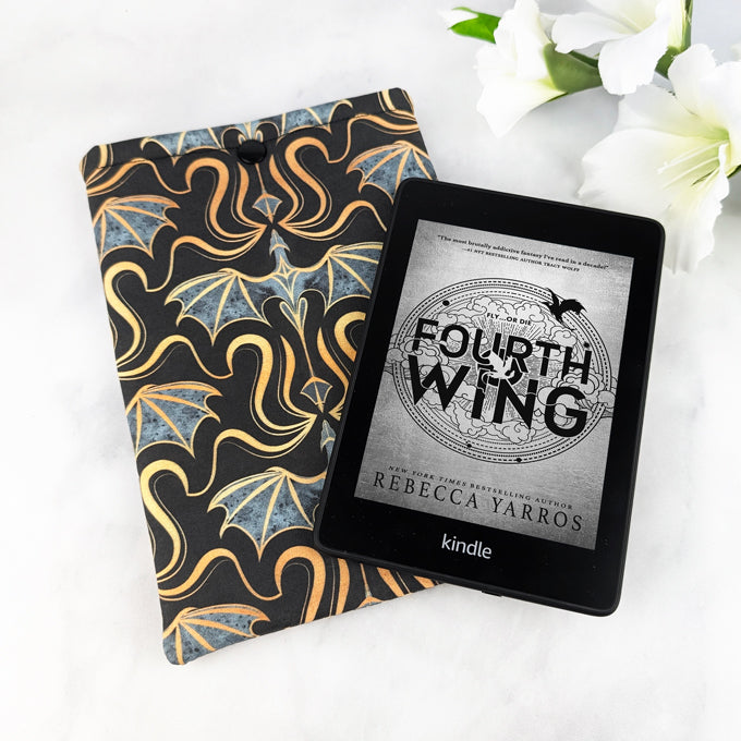 Padded Kindle Paperwhite sleeve with a bold pattern of gold and gray dragons on a black background, displayed with white flowers on a white surface.