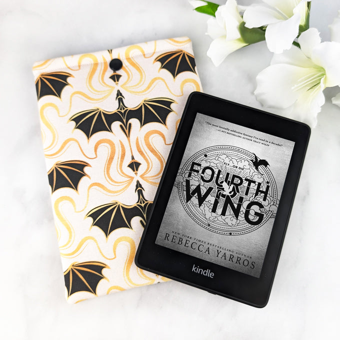padded kindle paperwhite sleeve with snap. fabric showcases simple illustrations of flying dragons in a damask style in shades of black, gold and white. Next to it there's a Kindle reader with Fourth Wing book cover on it.