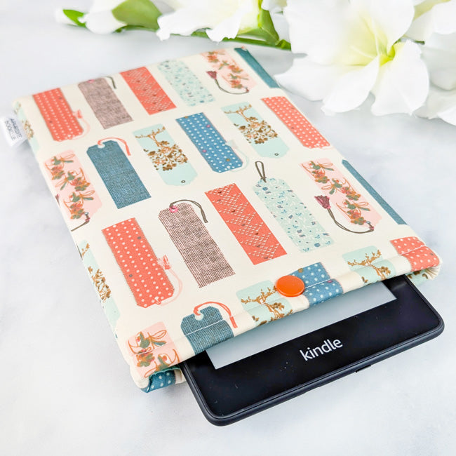 Kindle Paperwhite sleeve featuring a whimsical pattern of vintage-style bookmarks in coral, teal, and soft brown tones on a cream background.