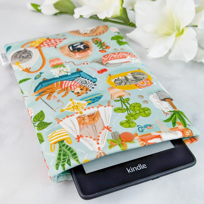 Kindle Paperwhite sleeve featuring a playful print of cozy lounging cats in beds, hammocks, and tents, surrounded by houseplants on a soft blue background.