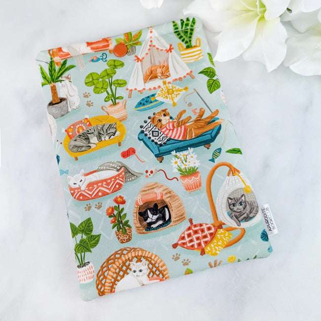 Kindle Paperwhite sleeve featuring a playful print of cozy lounging cats in beds, hammocks, and tents, surrounded by houseplants on a soft blue background.