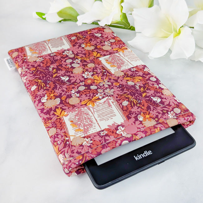 Fabric Kindle sleeve with a burgundy and pink floral pattern featuring open books, holding a Kindle device, displayed on a white surface with white flowers.