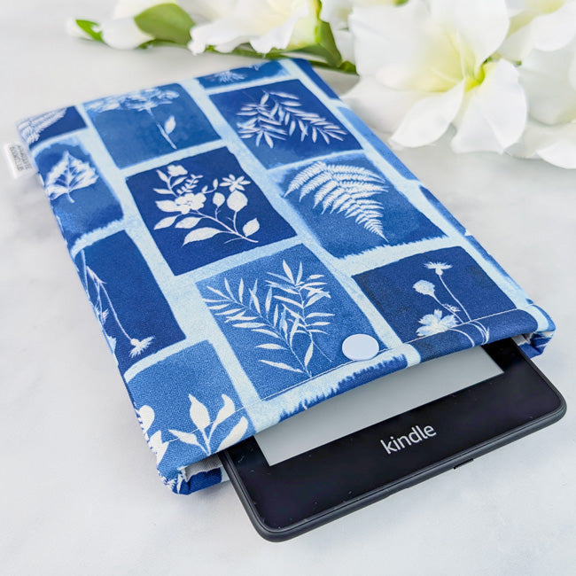 Kindle sleeve with a deep blue and indigo fabric featuring white botanical print blocks, shown with a Kindle inside and white flowers in the background.