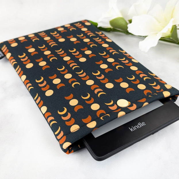 padded kindle paperwhite sleeve with snap. fabric showcases moon phases in beige and orange shades on dark navy background