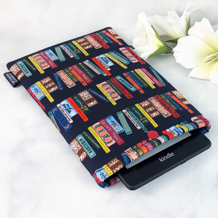 Handmade e-reader sleeve made from navy cotton fabric featuring colorful rows of illustrated classic book spines, including titles like Heidi and Gulliver’s Travels. The padded sleeve is shown holding a Kindle Paperwhite, displayed on a white marble background with white flowers for decoration.