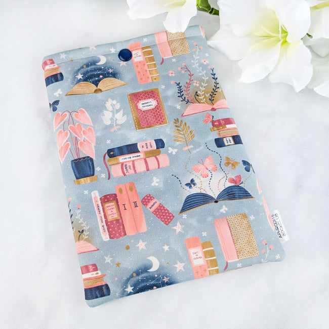 Fabric book sleeve featuring a whimsical print of pastel books, plants, and starry night scenes on a dusty blue background.