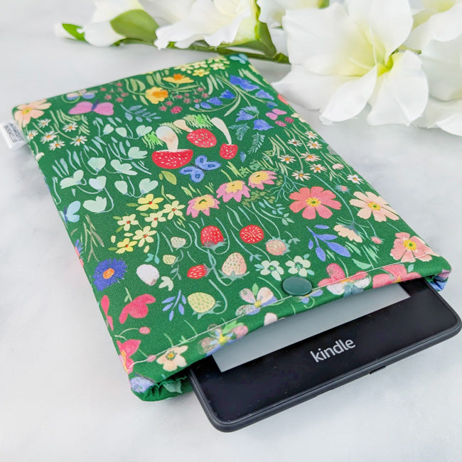 Kindle Paperwhite sleeve with a whimsical meadow design featuring colorful wildflowers, strawberries, and greenery on a rich green background.