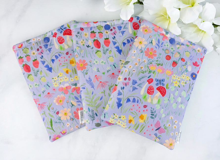 Three Kindle Paperwhite sleeves with a whimsical botanical print on lavender cotton, featuring strawberries, mushrooms, wildflowers, and fluttering butterflies.