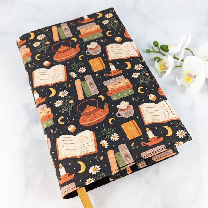 adjustable fabric book cover, fabric pattern shows teapots, book stacks, chamomile flowers, candles, moons and tea cups.