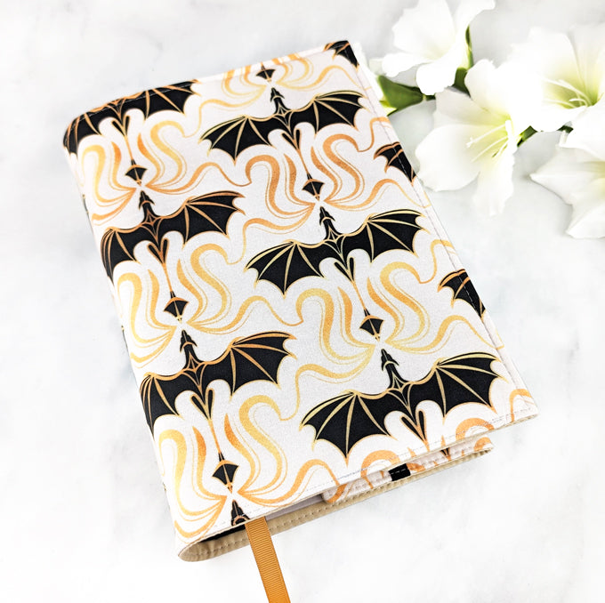 Fabric book cover with a bold pattern of gold and black dragons on a cream background, displayed with white flowers on a white surface.
