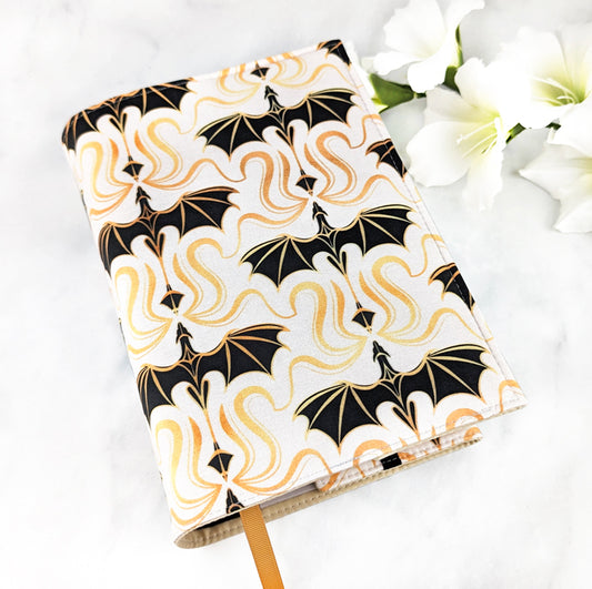 Fabric book cover with a bold pattern of gold and black dragons on a cream background, displayed with white flowers on a white surface.