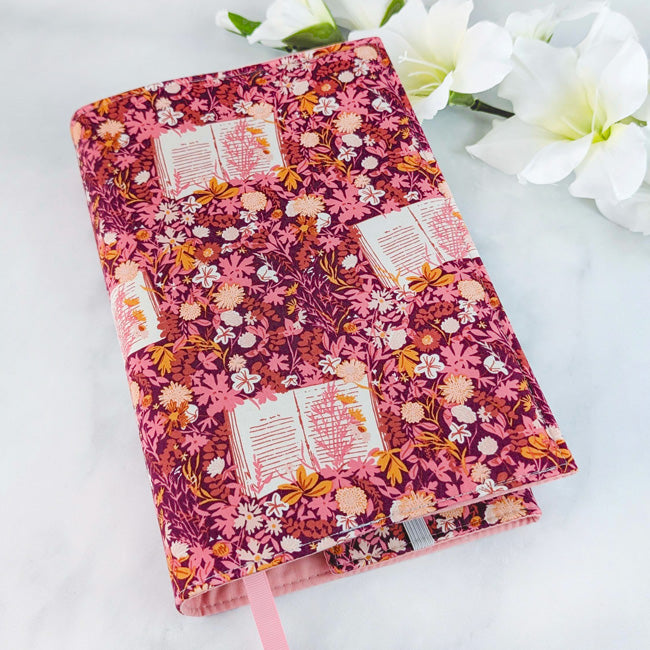 Fabric book cover with a mauve background and a whimsical print of open books, pink and peach flowers, and leafy accents. Finished with a soft pink ribbon bookmark.