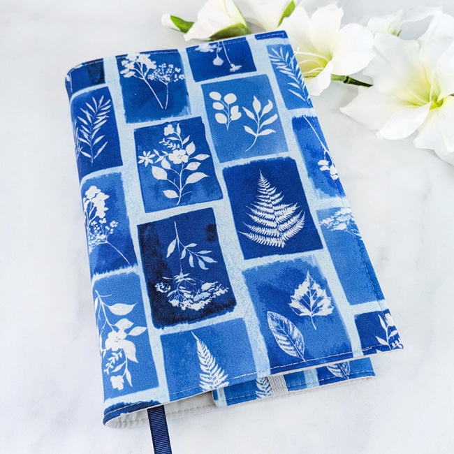 Fabric book cover with a blue and white botanical print resembling cyanotype art, featuring various leaves and flowers in rectangular frames, styled with white flowers.