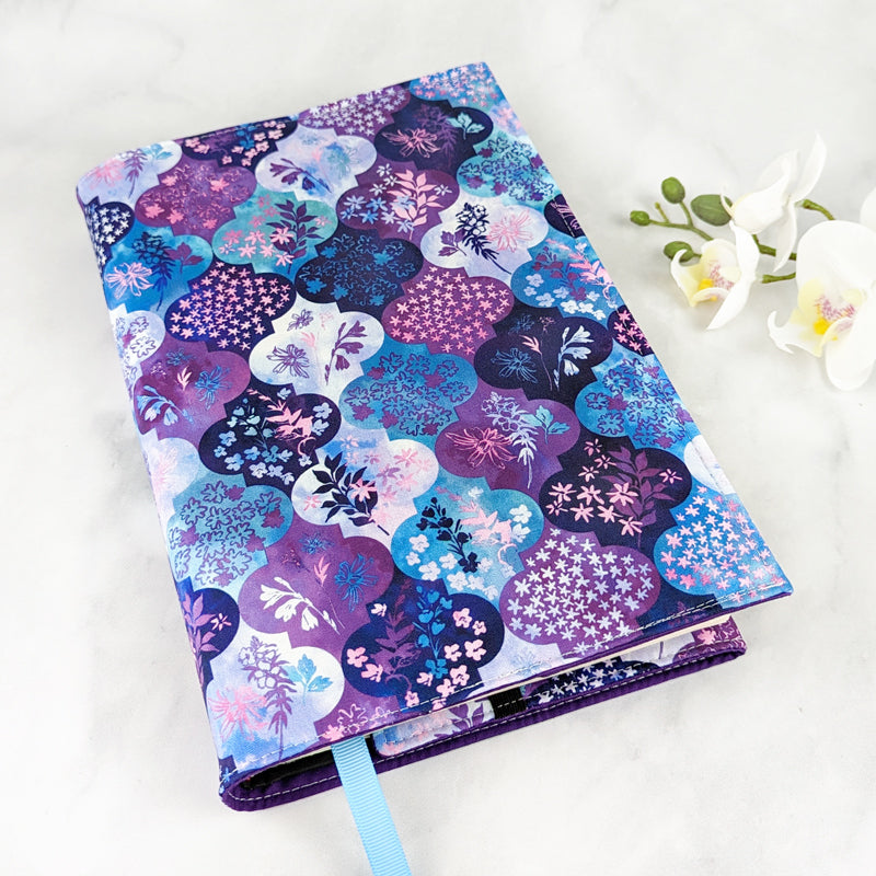 Fabric book cover with a vibrant geometric pattern of rounded tiles in shades of purple, blue, and teal, each featuring delicate floral motifs, finished with a light blue ribbon bookmark.