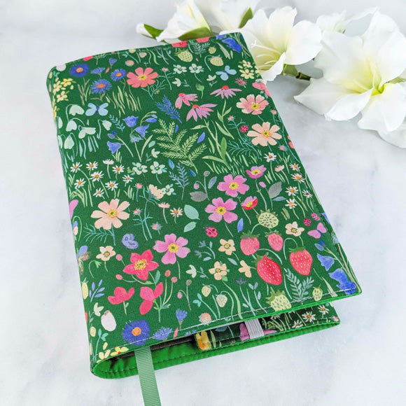 Alt text: Fabric book cover with a green background featuring a colorful wildflower and strawberry pattern, fitted on a book with a green ribbon bookmark, displayed beside white flowers.