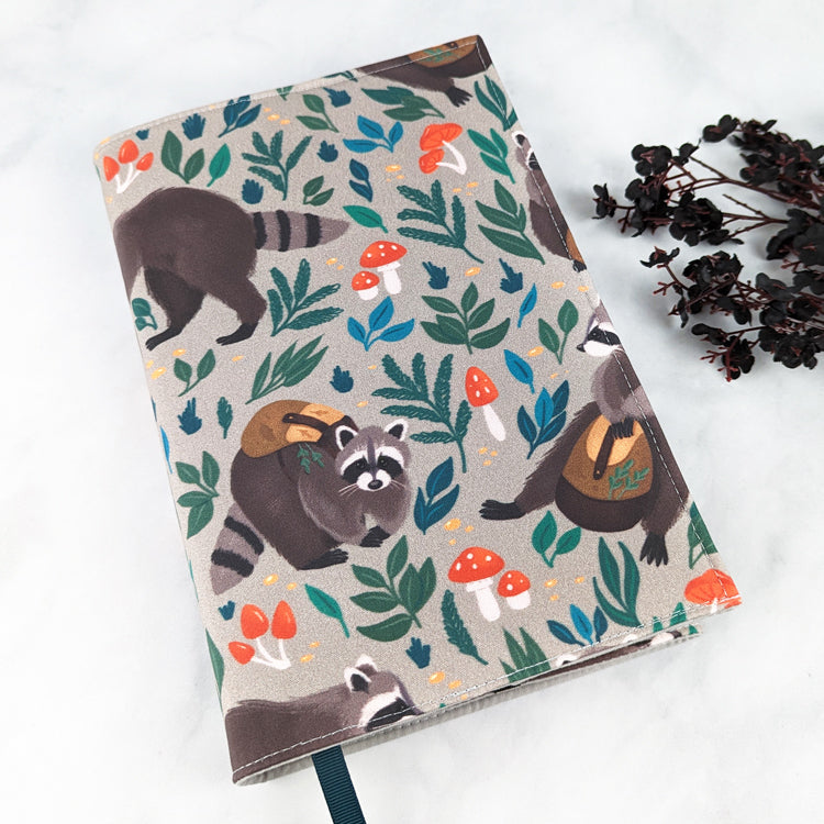 Fabric book cover featuring a playful woodland design with illustrated raccoons wearing backpacks, surrounded by leaves, mushrooms, and tiny flowers on a light gray background, finished with a dark teal ribbon bookmark.