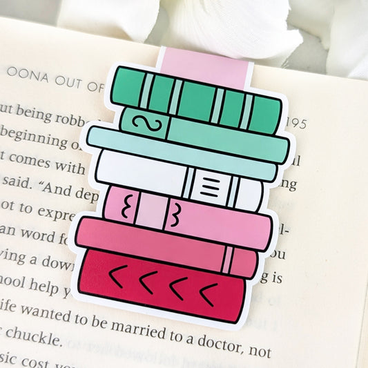 Magnetic bookmark shaped like a stack of books in abrosexual pride colors—shades of green, white, and pink—clipped to the top of an open book.