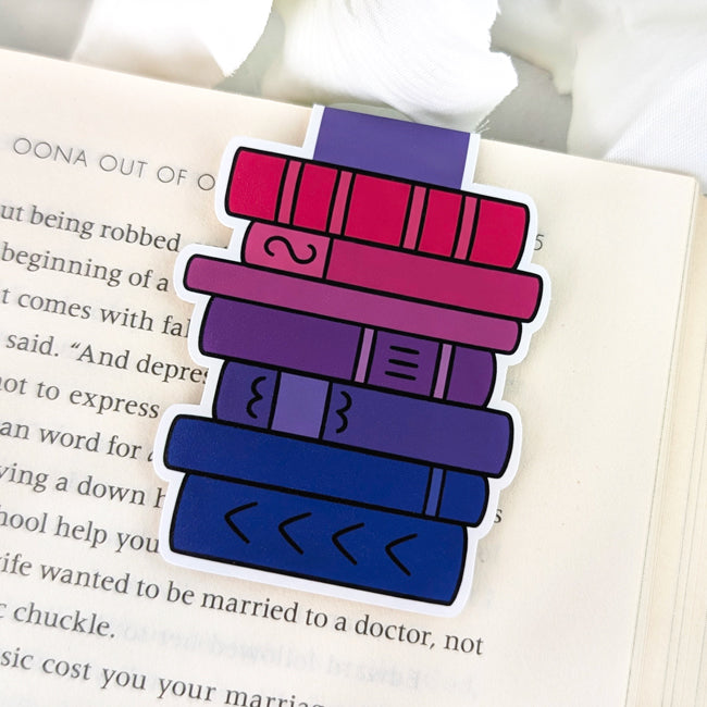 Magnetic bookmark shaped like a stacked pile of books in bisexual pride flag colors—pink, purple, and blue—clipped to the top of an open book page.