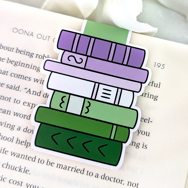 Magnetic bookmark shaped like a stack of books in genderqueer pride colors—purple, white, and green—clipped to the top of an open book page.
