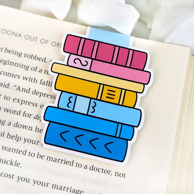 Magnetic bookmark shaped like a vertical stack of books, attached to the top of an open book. Each book spine is colored in pink, yellow, and blue, representing the pansexual Pride flag.