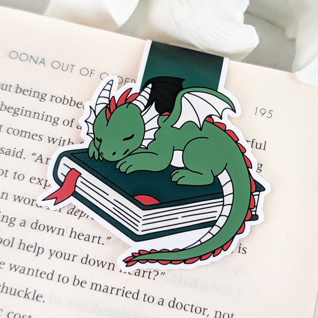 Magnetic bookmark featuring a cute green dragon with red accents sleeping on a closed book with a red ribbon bookmark.