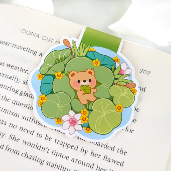 Magnetic bookmark featuring a cute bear holding a frog while sitting among lily pads and flowers in a pond, clipped to the top of an open book.