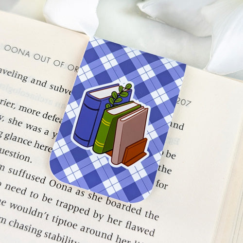 Magnetic bookmark featuring a stack of colorful books with a sprig of leaves, set against a blue argyle background, clipped to the top of an open book.