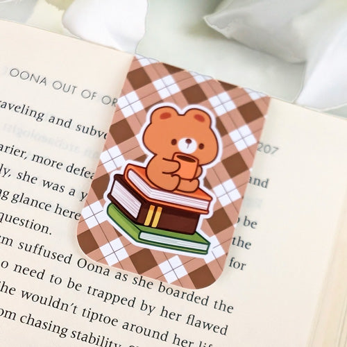 Magnetic bookmark featuring a cute brown bear holding a mug while sitting on a stack of books, set against a brown argyle background, clipped to the top of an open book.