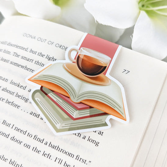 Magnetic bookmark with coffee cup on stack of books, clipped to an open book with white flowers nearby.