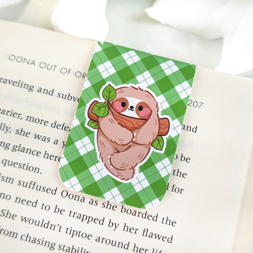 Magnetic bookmark featuring a cute sloth hugging a branch with leaves, set against a green argyle background, clipped to the top of an open book.