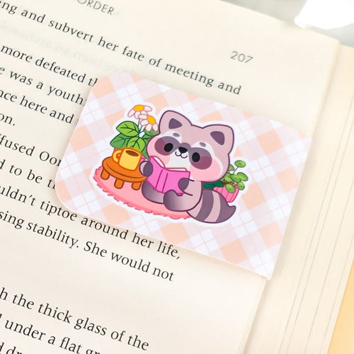 Magnetic bookmark featuring a cute raccoon reading a book next to a teacup and potted plant, set against a peach argyle background, clipped to the top of an open book.