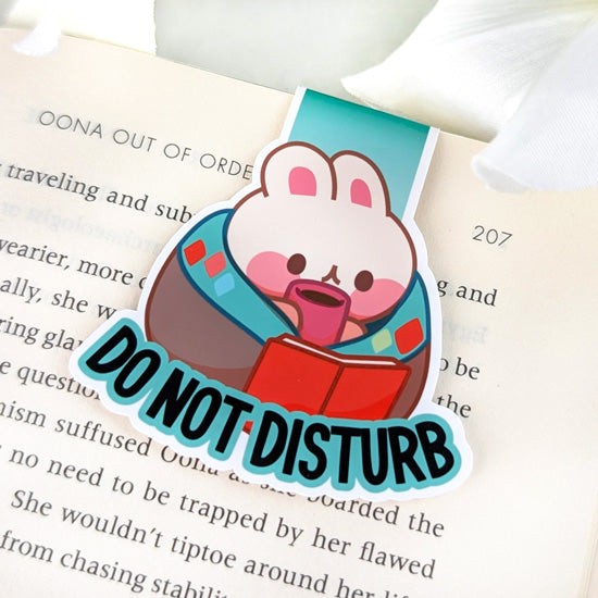 Magnetic bookmark featuring a bunny wrapped in a blanket, holding a book and a mug, with the text “DO NOT DISTURB,” clipped to the top of an open book.
