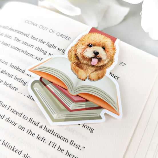 Magnetic bookmark featuring a fluffy goldendoodle puppy sitting on an open book, clipped to a page with white flowers nearby.
