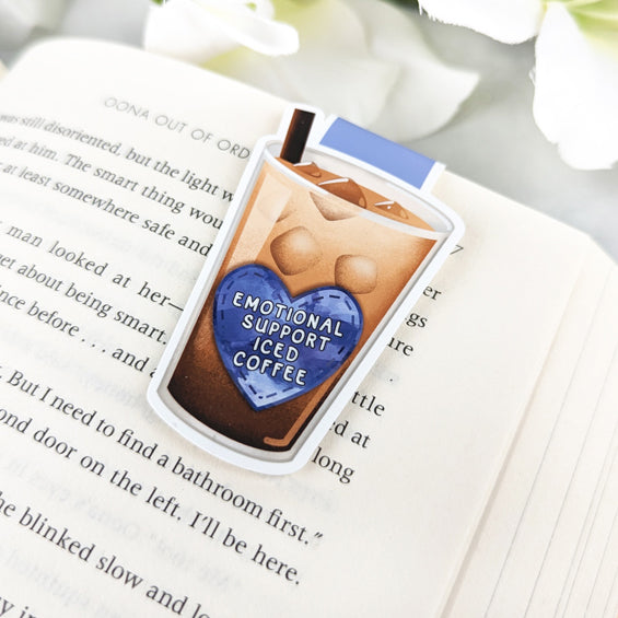 Magnetic bookmark shaped like a glass of iced coffee with a blue heart that reads 'Emotional Support Iced Coffee,' clipped to an open book.