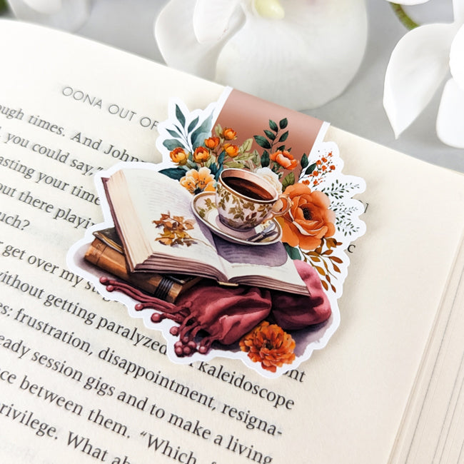Fall-themed magnetic bookmark featuring a vintage teacup, open book, orange flowers, and cozy autumn accents, clipped to an open book.